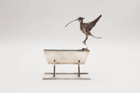  Paliteiro em prata representando um pássaro de asas abertas junto a bebedouro em movimento.